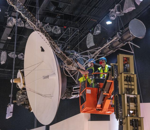 Two men in yellow construction vests and helmets use a lift to install the antenna of a spacecraft