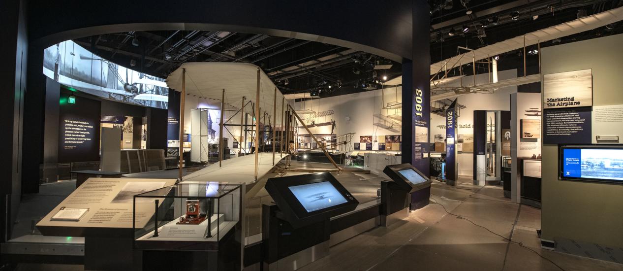 A view of the Wright Brothers gallery, with the Wright Flyer in the center surrounded by display panels and touch screens. 