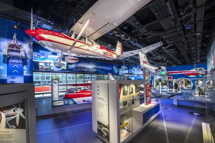 A view of the Thomas W. Haas We All Fly Gallery, showing cases on the ground under a suspended red and white airplane. 