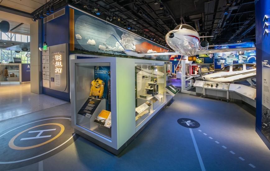 A view of the We All Fly Gallery. A large airplane can be seen hanging overhead and a case with a jacket in it viewable from the ground. 