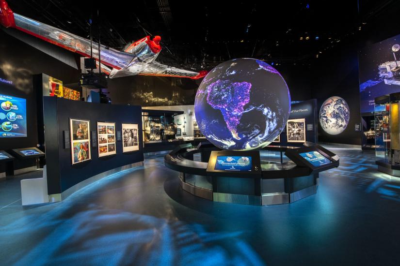 A view of the One World Connected gallery. There is a large glowing globe visible at the center of the room, images around it, and a silver and red plane hanging overhead. 