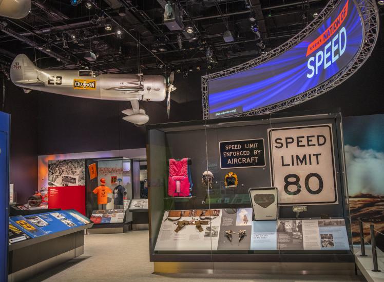 A photo of the inside of the Nation of Speed exhibit. A sign that says Nation of Speed hangs in the top right corner and across from it is Roscoe Turners airplane. A case with side signs that say speed limit 80 and speed limit enforced by aircraft is in the foreground.