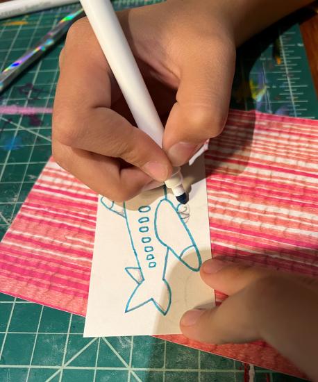 A child's hand draws a plane on a rectangular piece of paper. 