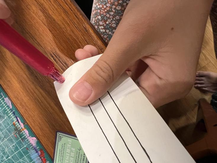 A craft activity in process for Soar Together. Hands hold a small implement up to a rectangular sheet with lines on it. 