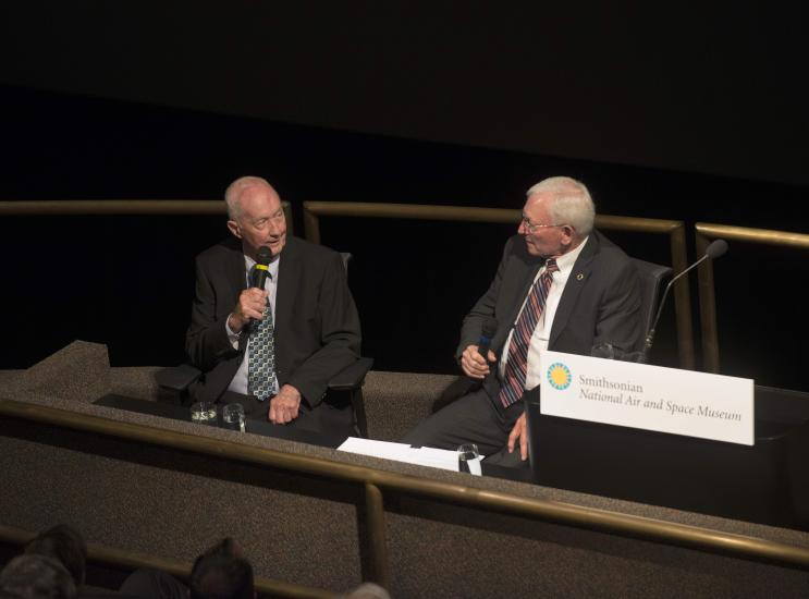 Two men sitting in chair with business suits on and microphones in their hands.