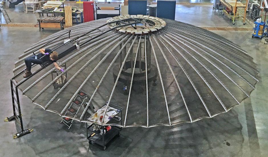 A person lying on top of a large umbrella shaped satellite antenna while cleaning it.