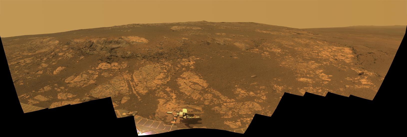 A panoramic view looking out over a hill of the dusty red terrain on Mars.