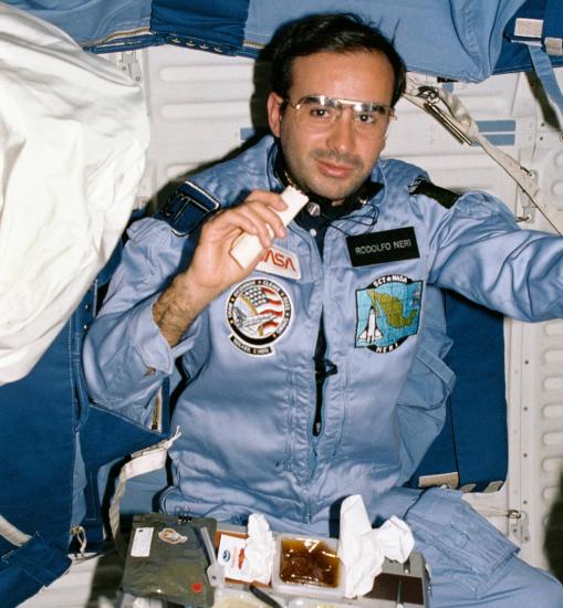 An astronaut in a blue suit floats inside a spacecraft. His food tray is on his lap, a tortilla in his hand. 