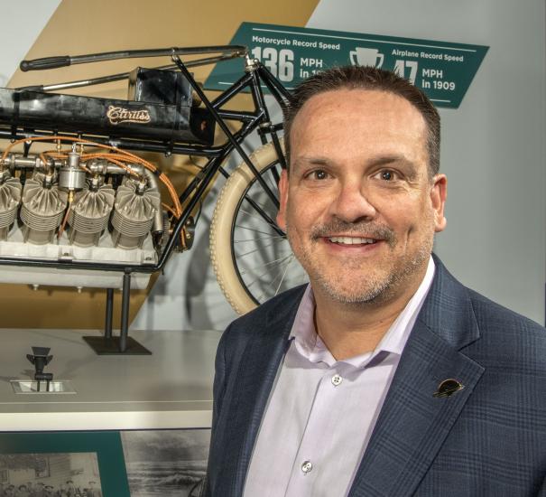 A man in a blazer smiles in front of the camera in front of a Curtiss bicycle. 