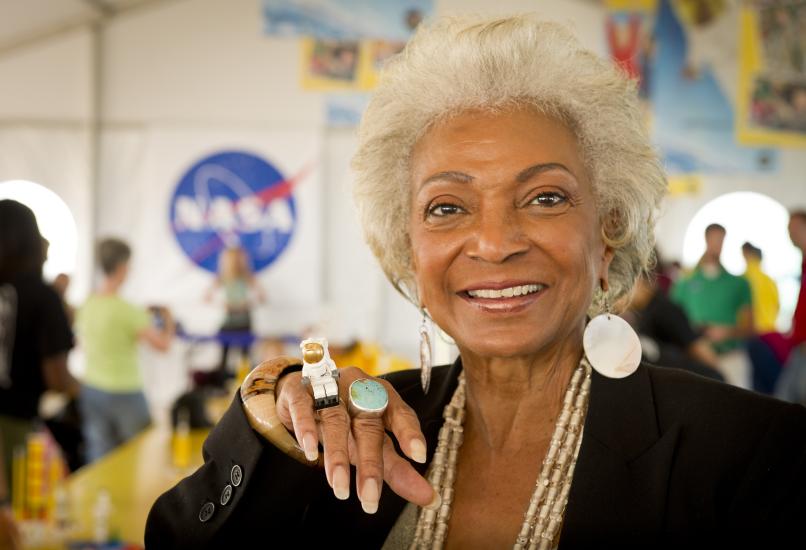 Nichelle Nichols, later in her life, smiles for a photo with a small LEGO mini figure resting on her hand and the NASA logo in the background. 