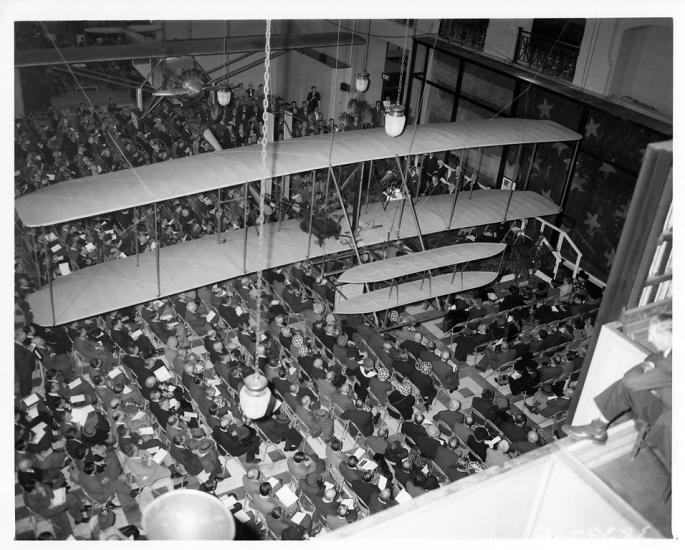 A photo taken from a balcony looking down at a crowd of seated people. The Wright Flyer hangs overs head, with the Spirit of St. Louis hung across from it.