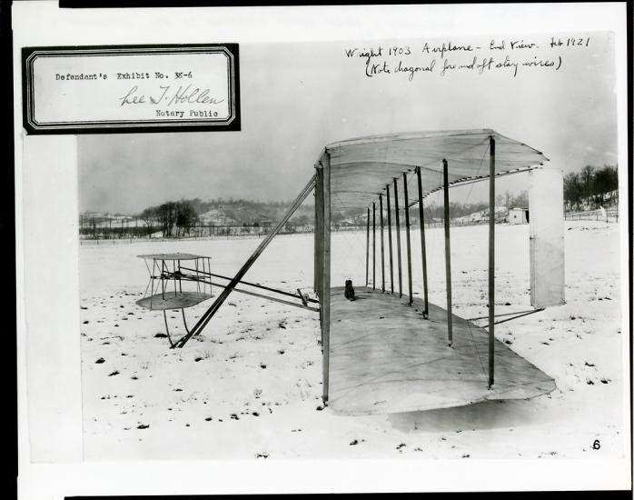 The Wright Flyer as seen from the side in a snowy field. There is an exhibit number stamped at the top left.