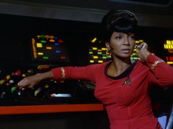 A Black woman in a red starfleet uniform looks up from a dashboard. 