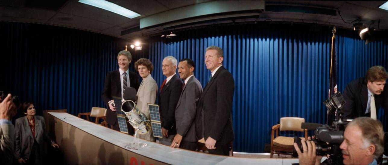 A group of 5 people stand shoulder to shoulder in front of a model of a telescope behind a table. On the other side of the table are members of the press.