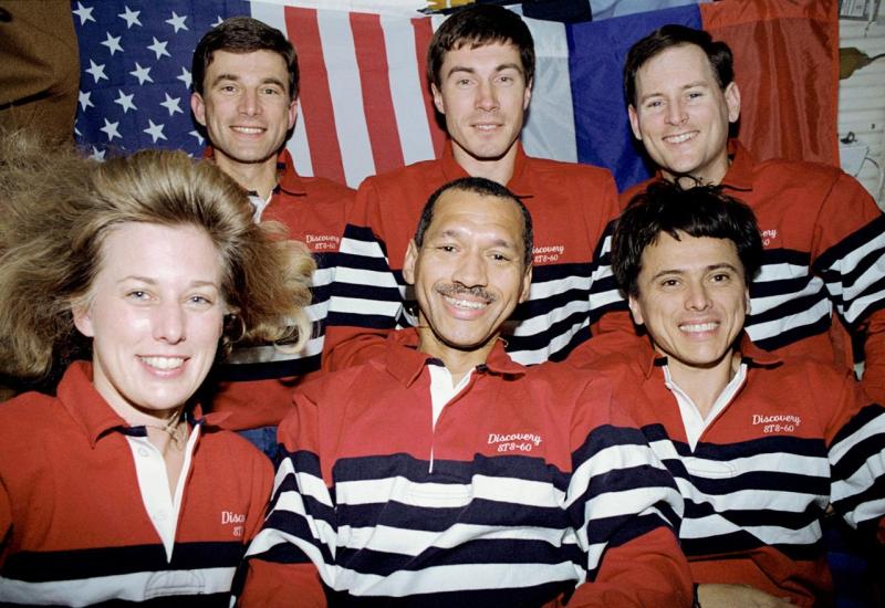 Six people pose in two rows of three aboard a space shuttle. The woman in the left demonstrates the weightlessness of space with her hair floating freely.