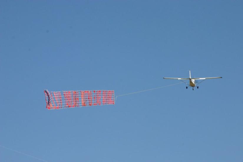 A plane, flying, carries a banner with red letters on it.