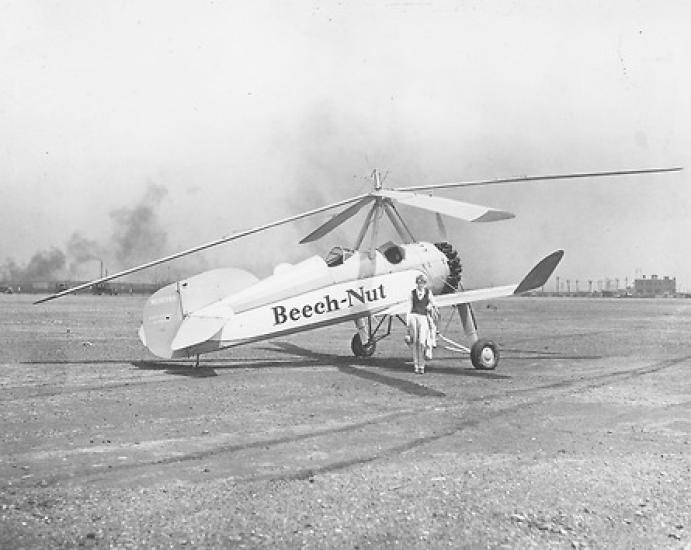 A black and white photograph of a woman standing next to an auto-giro that reads "Beech Nut" on the side.