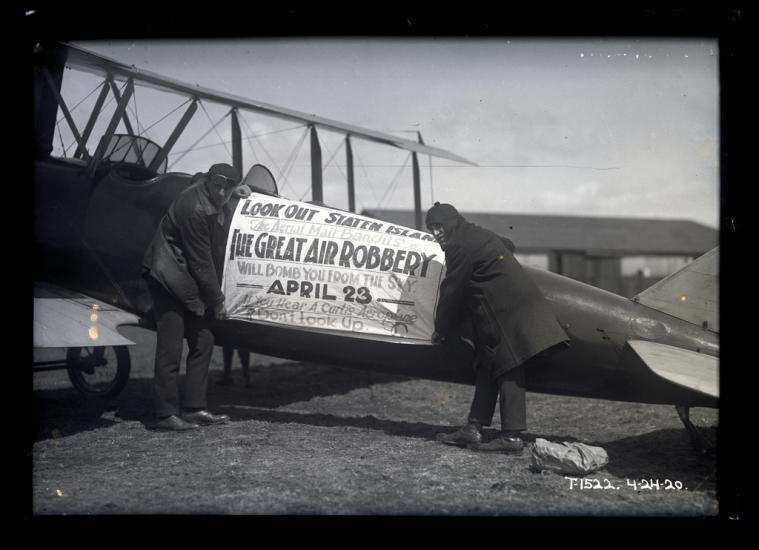 Men affix banner to side of biplane fuselage which reads; Look Out Staten Island / The Aerial Mail Bandits in / The Great Air Robbery / Will Bomb You From The Sky / April 23. If You Hear A Curtis [sic] Aeroplane / Don't Look Up.