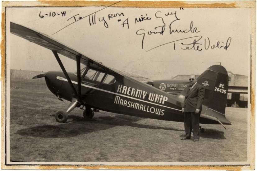 A sepia tone photograph of a small airplane that reads "Kreamy Whip Marshmallows" on the side. A man stands next to it.