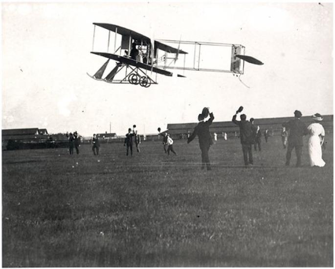 A black and white image of a biplane flying low to the ground. People on the ground wave their caps. 