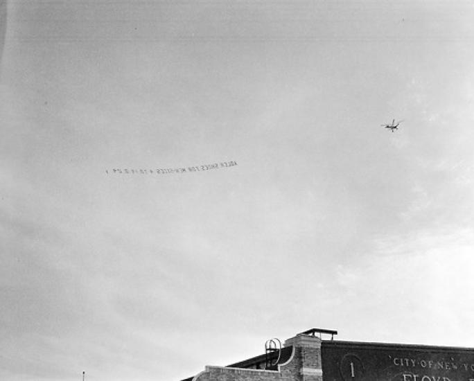 Distant right rear view of a Pitcairn PCA-2 Autogiro towing an aerial banner reading, "Adler Shoes for Men - sized 4 to 14 & 24."Due to the direction of flight, the banner reads in reverse.