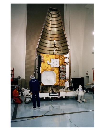 A large golden colored cube inside a metal conical encasing is surrounded by various technicians performing different activities on the structure.