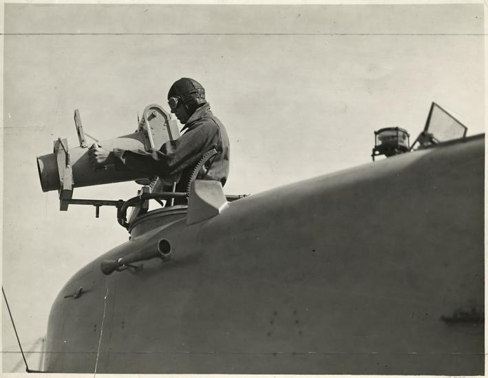 A close-up view from low left side of nose of aircraft, looking forward, of a man in a flight cap that stands in the forward cockpit of a Naval Aircraft Factory F-5L (PN-5) demonstrating a hand-operated K-2 aerial camera used for oblique aerial photography.