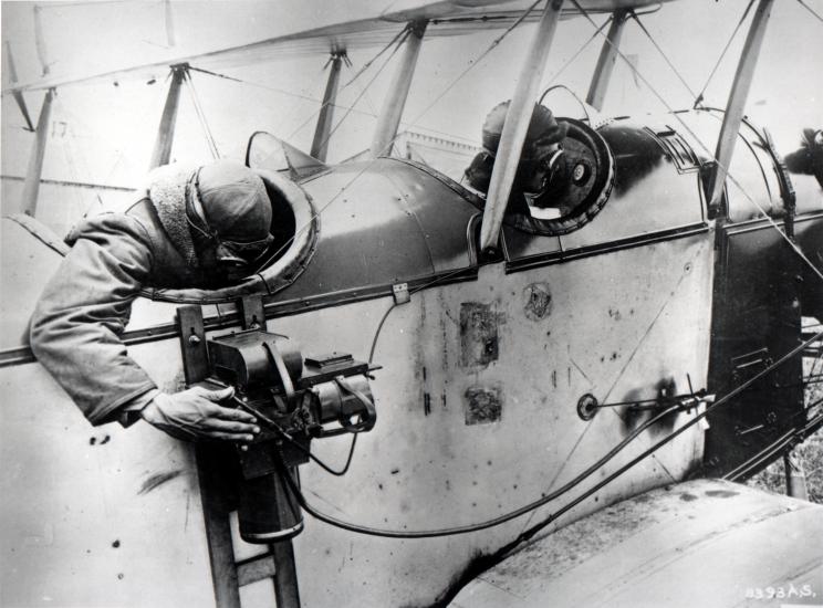 A man reaches out from the back seat of a biplane to look into the viewfinder of a camera that is strapped to the side of the aircraft.