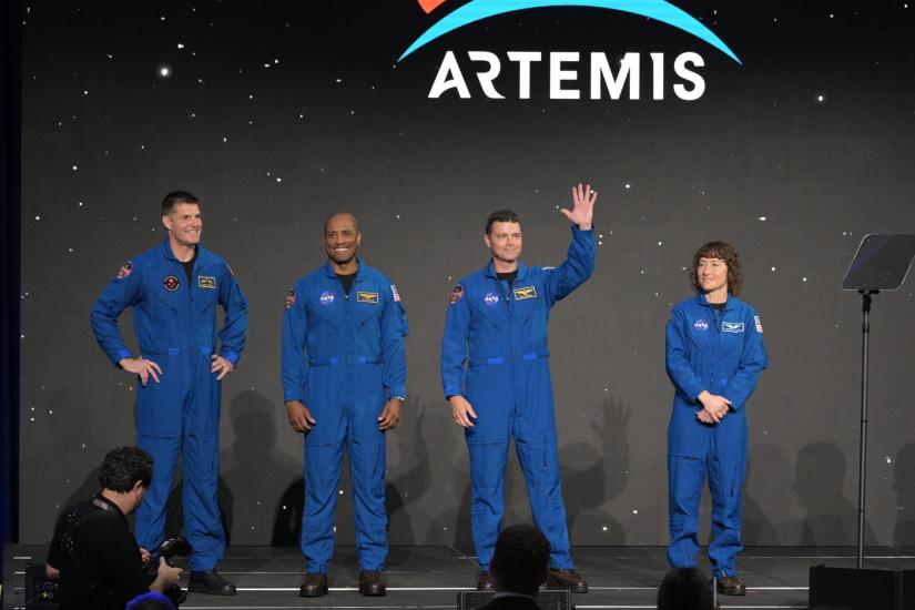 Left to right—astronaut Jeremy Hansen, Victor Glover, Reid Wiseman, and Christina Koch. All wearing blue flight suits standing on a stage. 