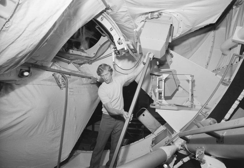 Black and white image of a man holding up a device in a space station simulator.