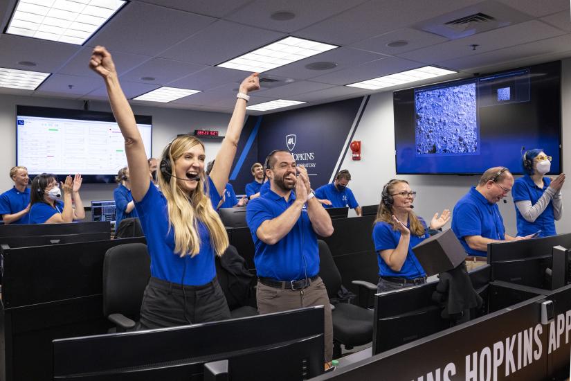 People in blue polo shirts standing in front of computer clap and raise their arms in celebration.