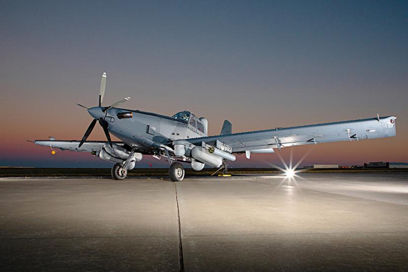 Low angle view of the OA-1K Sky Warden, a large grey airplane with two very large wings and a five blade propellor on the nose. The photographs is taken on tarmac at night