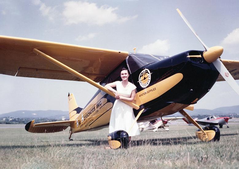 Woman in white dress poses next to a Cessna 140, a small black and yellow plane with a propeller, on grass runway.