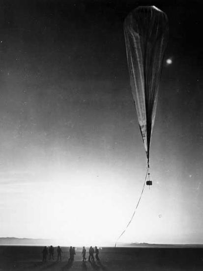 A balloon floats just above the Earth's surface with a camera attached to it. A group of people stand in a semi circle on the ground below it.