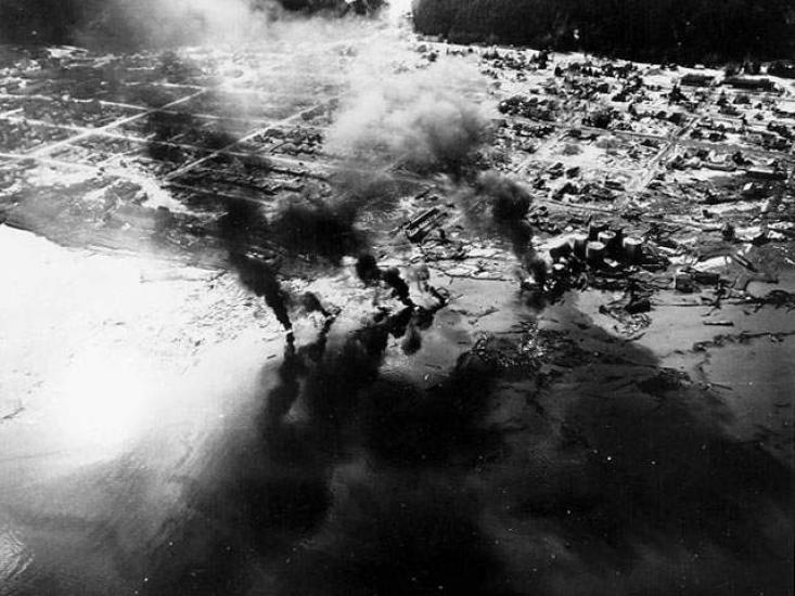 An aerial photograph of a small city with large plumes of smoke coming up.