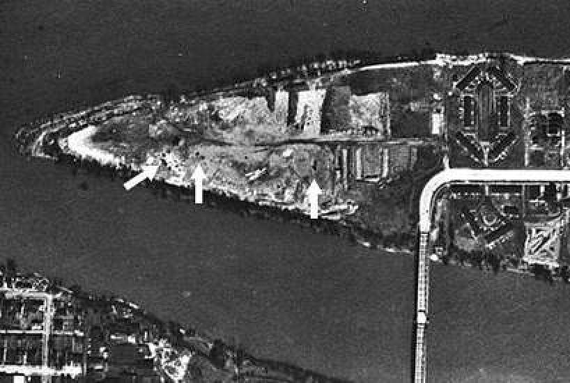 An aerial photo of a triangular shaped island that has a bridge connecting it to the mainland. Arrows point to dark patches on the southern parts of the island.