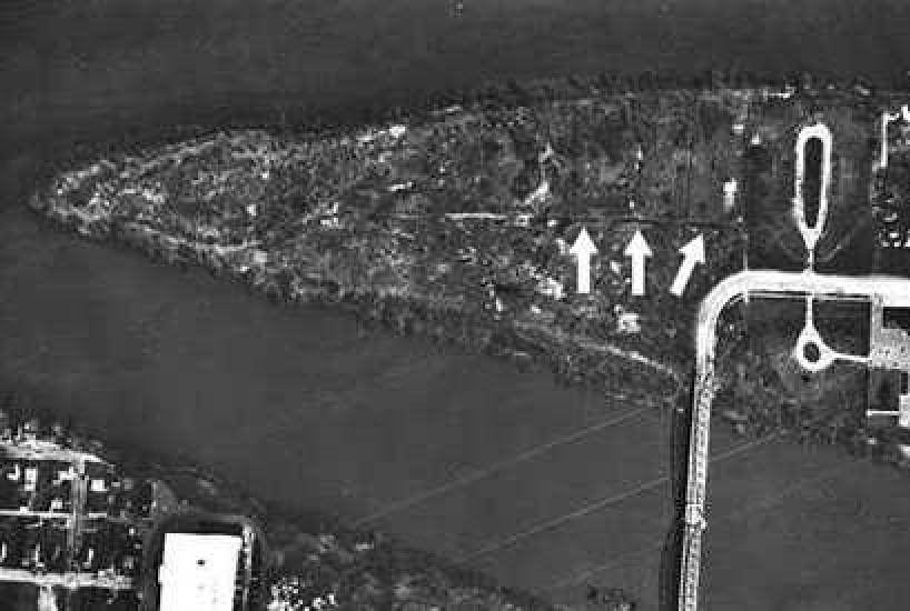 An aerial photo of a triangular shaped island that has a bridge connecting it to the mainland. Arrows point to dark spots at the center of the island.