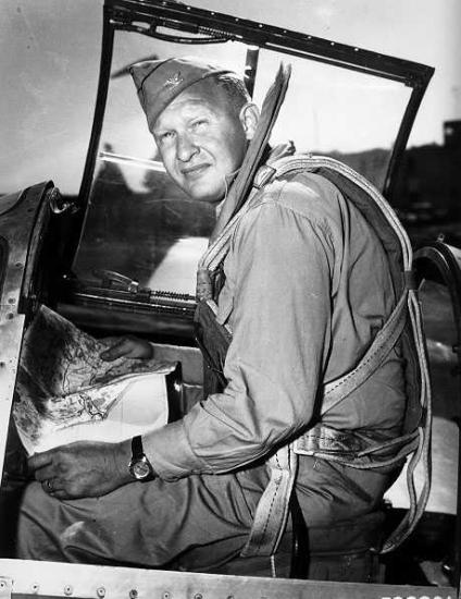 Photograph of a man sitting in an aircraft holding a map and looking toward the camera.