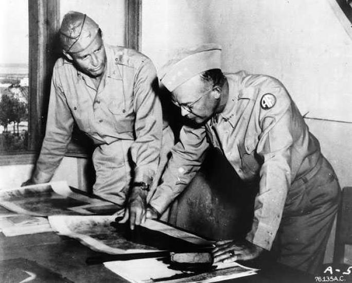 Two men in military uniforms stand at a table looking at photographs.