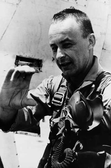 A man in a flight suit turned three-quarters of the way toward the camera, he makes a c-shape with his hand.