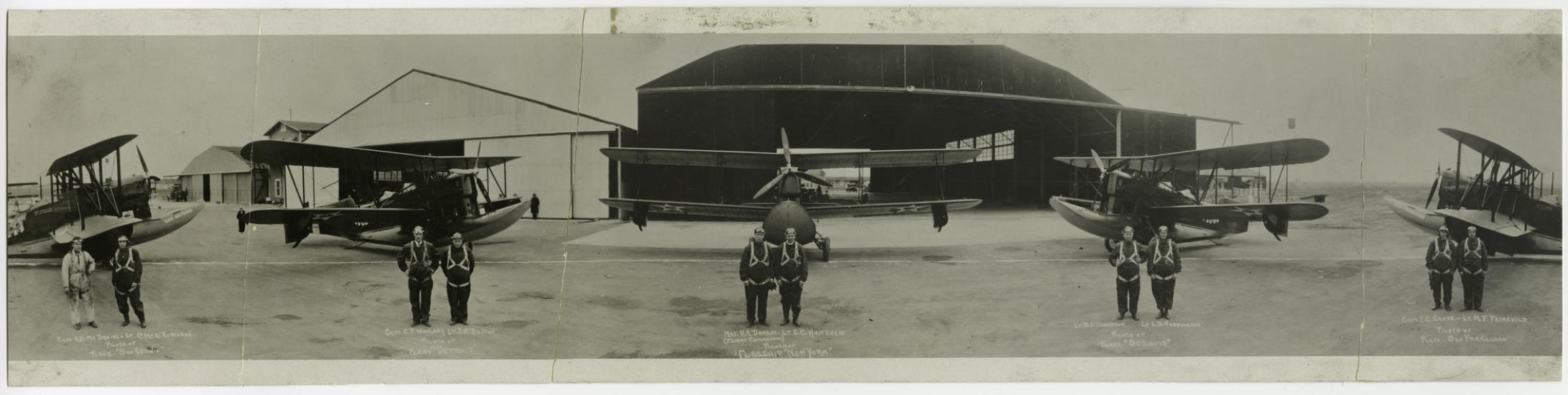 Black and white panoramic photograph of five airplanes, each plane has two men in flightsuits standing in front, with a hangar in the background. There are folds throughout the image.