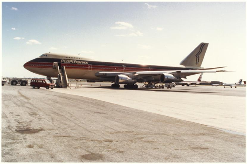 Color photograph of jet aircraft with red white and blue stripes and the text "PeoplExpress" on the fuselage. A stylized drawing of two parallel faces in profile are on the tail.