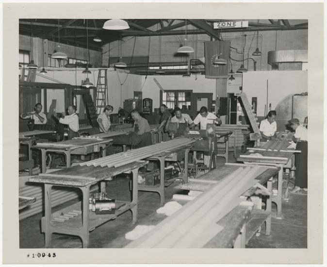 Black and white photograph of corrugated metal pieces with men and women working in the background