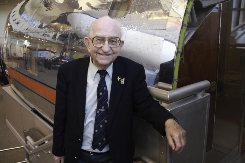 Balding elderly man wearing glasses stands beside a silver reflective airplane nose.