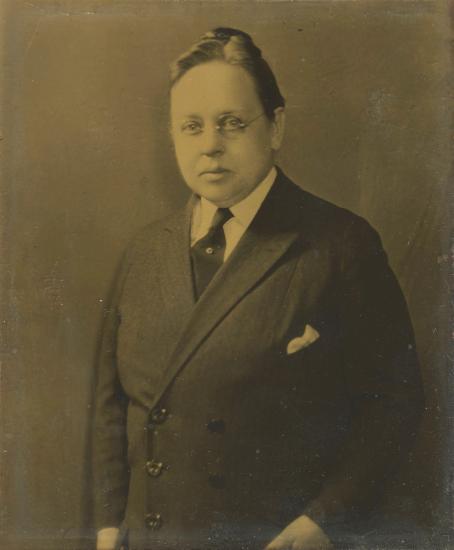 Sepia-toned photo of a woman wearing white collar, dark Windsor tie held with a stick pin, and a double breasted coat