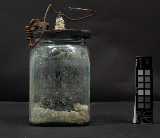 An early battery that resembles a jar with substance inside is photographed against a dark background.