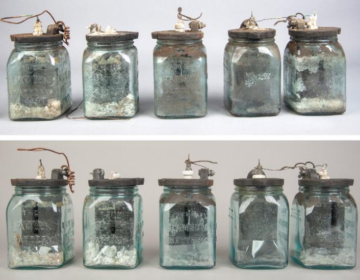 Five early batteries that resembles a jar are photographed and compared. The bottom set of batteries appear cleaner.