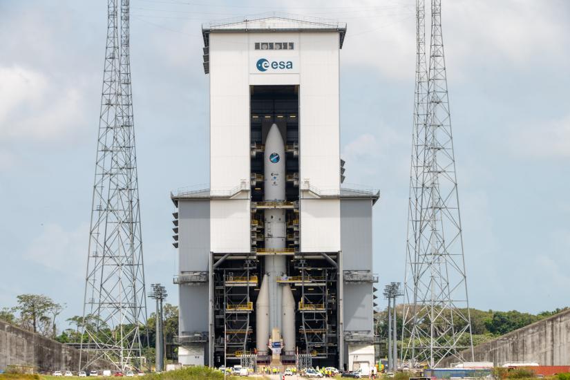 Ariane 6 rocket inside a large building. There are large cut-out doors and the rocket is photographed through them from the outside of the building.