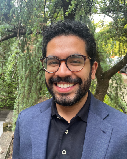 Portrait of a Mexican-American man. He is wearing a suit and smiling and there is a tree behind him.