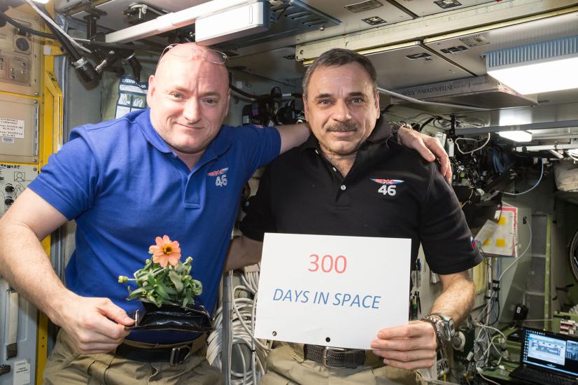 Two astronauts float on the International Space Station. One wears a blue polo and holds a flower, the other wears a darker polo and holds a sign that says '300 Days in Space.'